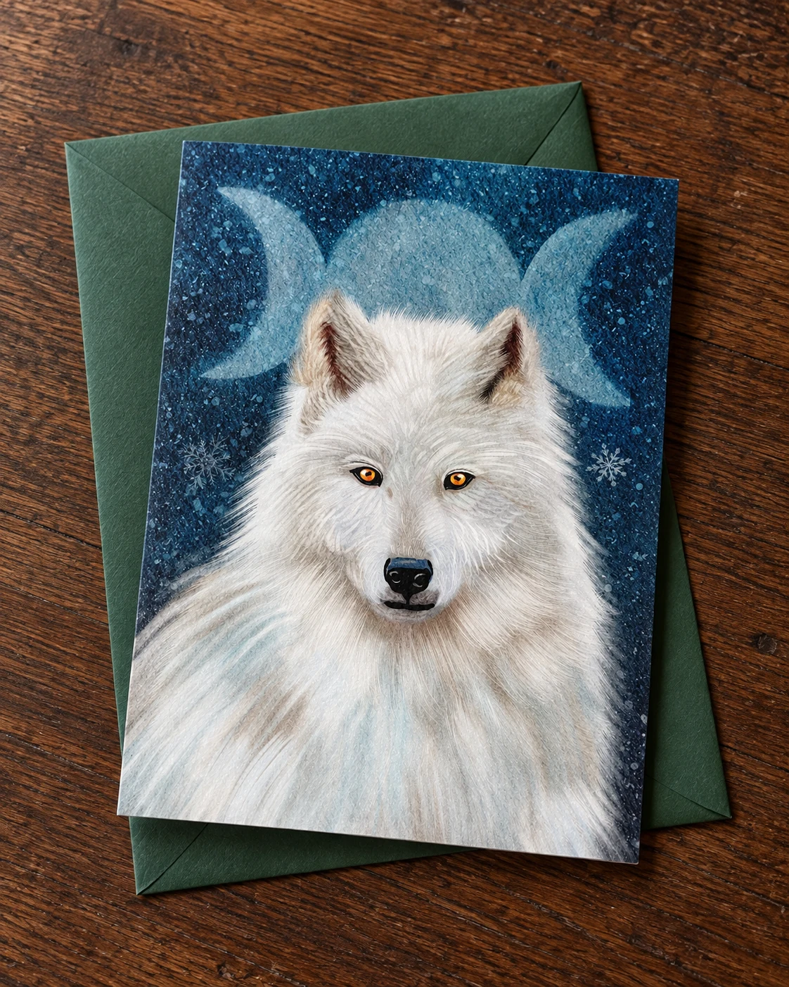 Beneath the Three Moons borderless 5 × 7 inch greeting card on a green envelope on an oak desk